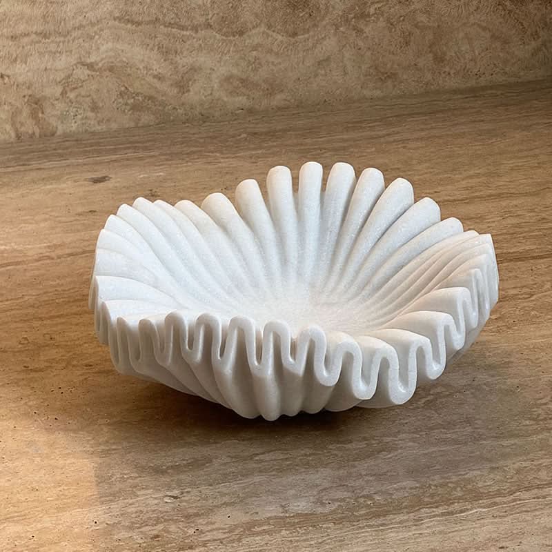 White fluted marble fruit bowl with a wavy edge, placed on a beige stone surface.