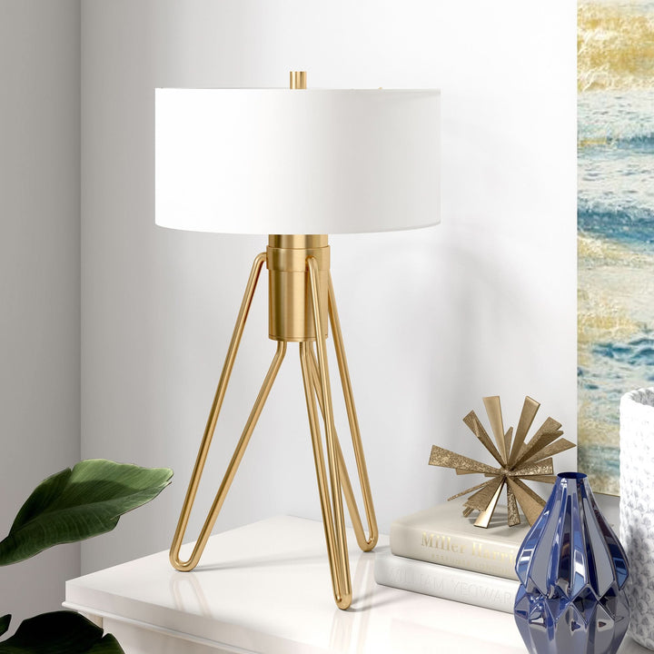 Brass tripod mid-century table lamp with white drum shade on a white table.