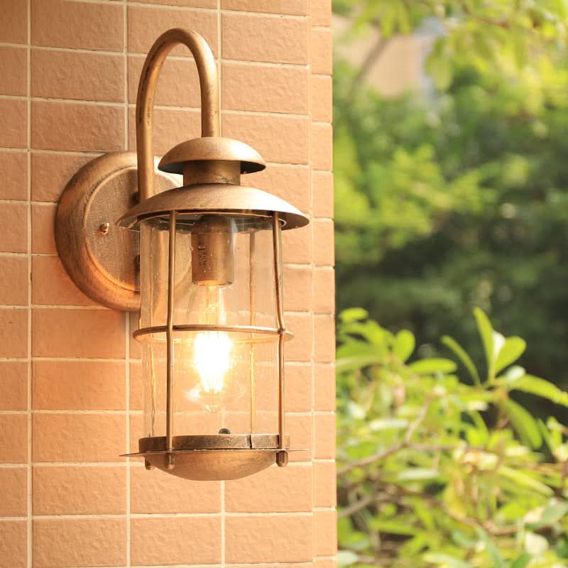 Rustic bronze cylindrical outdoor sconce with glass shade on tan tiled wall.