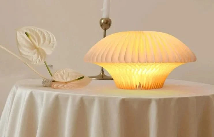 Origami bell-shaped paper bedside lamp glows warmly on a table with white anemone flowers.