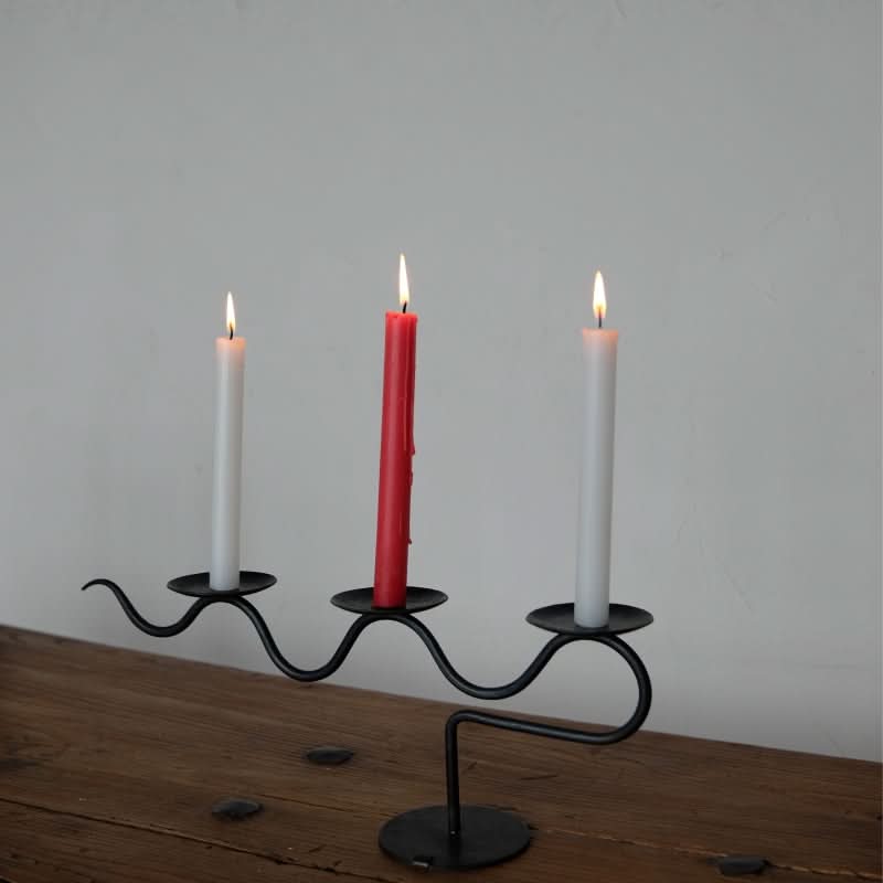 Black metal wavy three-candle holder with red and white candles on a wooden table.