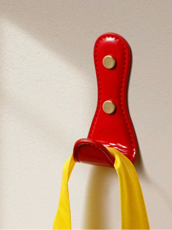 Red leather wall hook with brass accents, holding a yellow strap. Dopamine Decor.