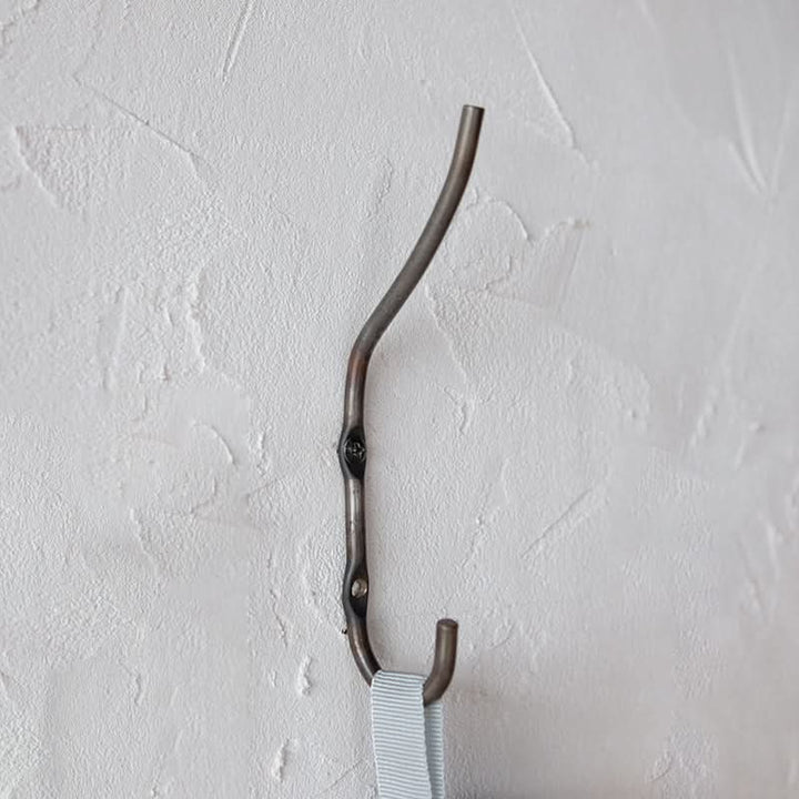 Rustic dark metal Industrial Style Wall Hook with light blue striped strap hanging from it on a textured white wall.