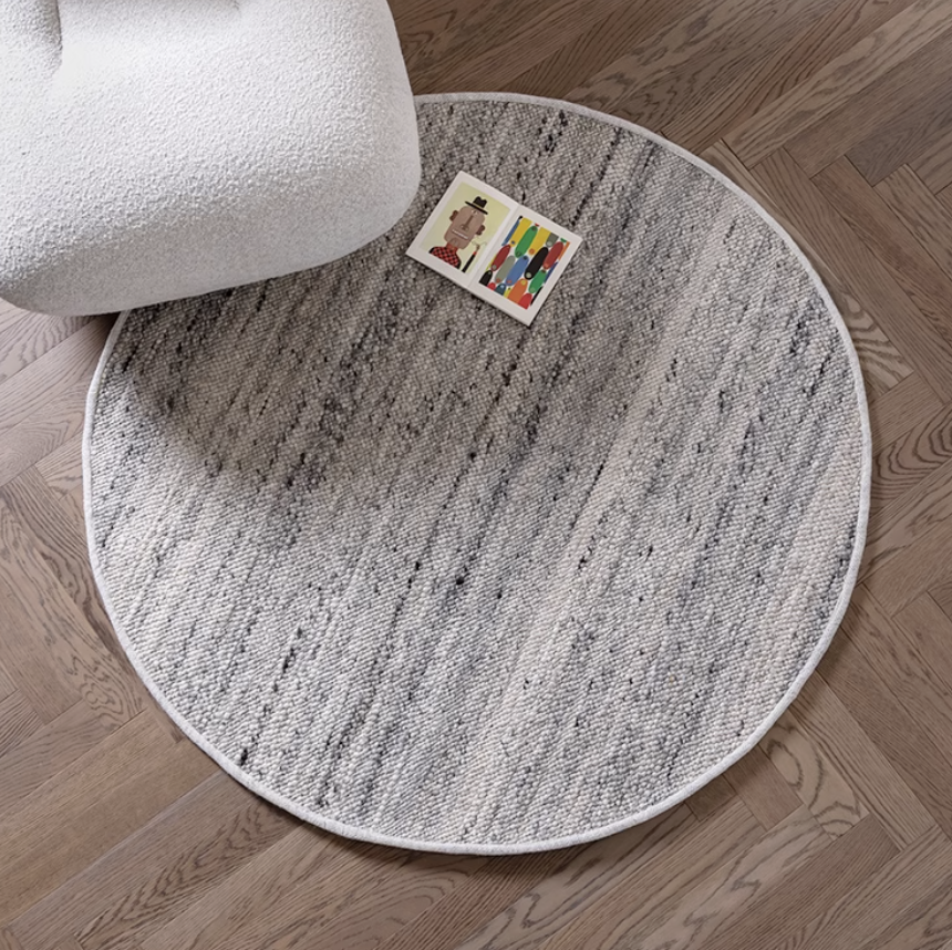 Round natural wool carpet in grey and white tones on a wood floor, near a white armchair.