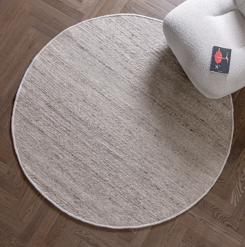 Round natural wool carpet in neutral tones, placed on a wooden floor next to a cream armchair.