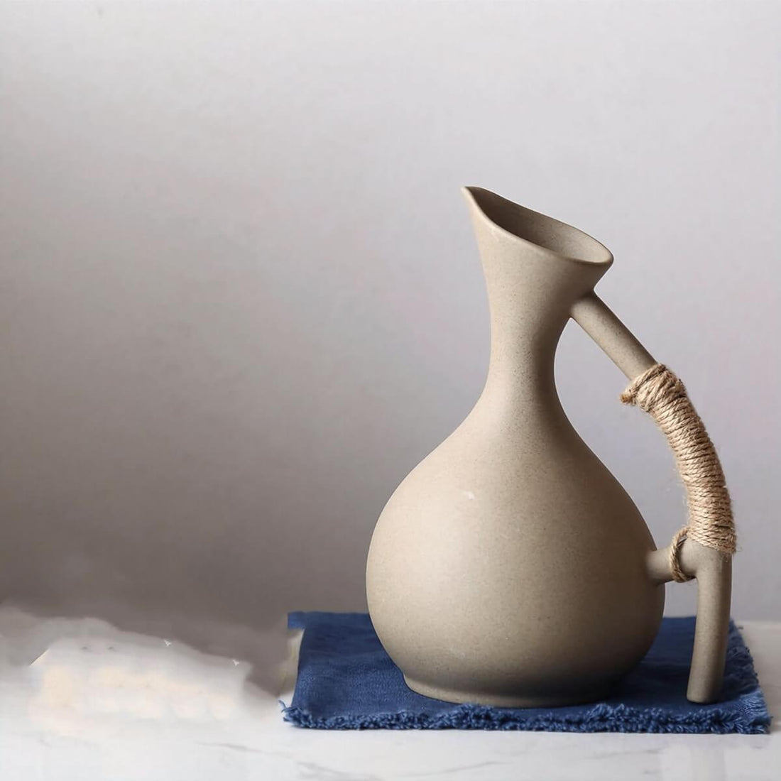 Abstract ceramic pitcher with arched handle, natural tone, on blue cloth. Home decor.