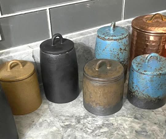 Rustic ceramic tea canisters in blue, bronze, and black finishes, with looped handles, on a countertop.