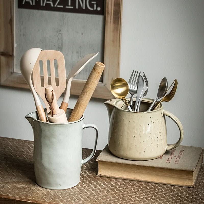 Farmhouse ceramic pitcher utensil holder with kitchen forks, spoons, and spatulas on a rustic table.