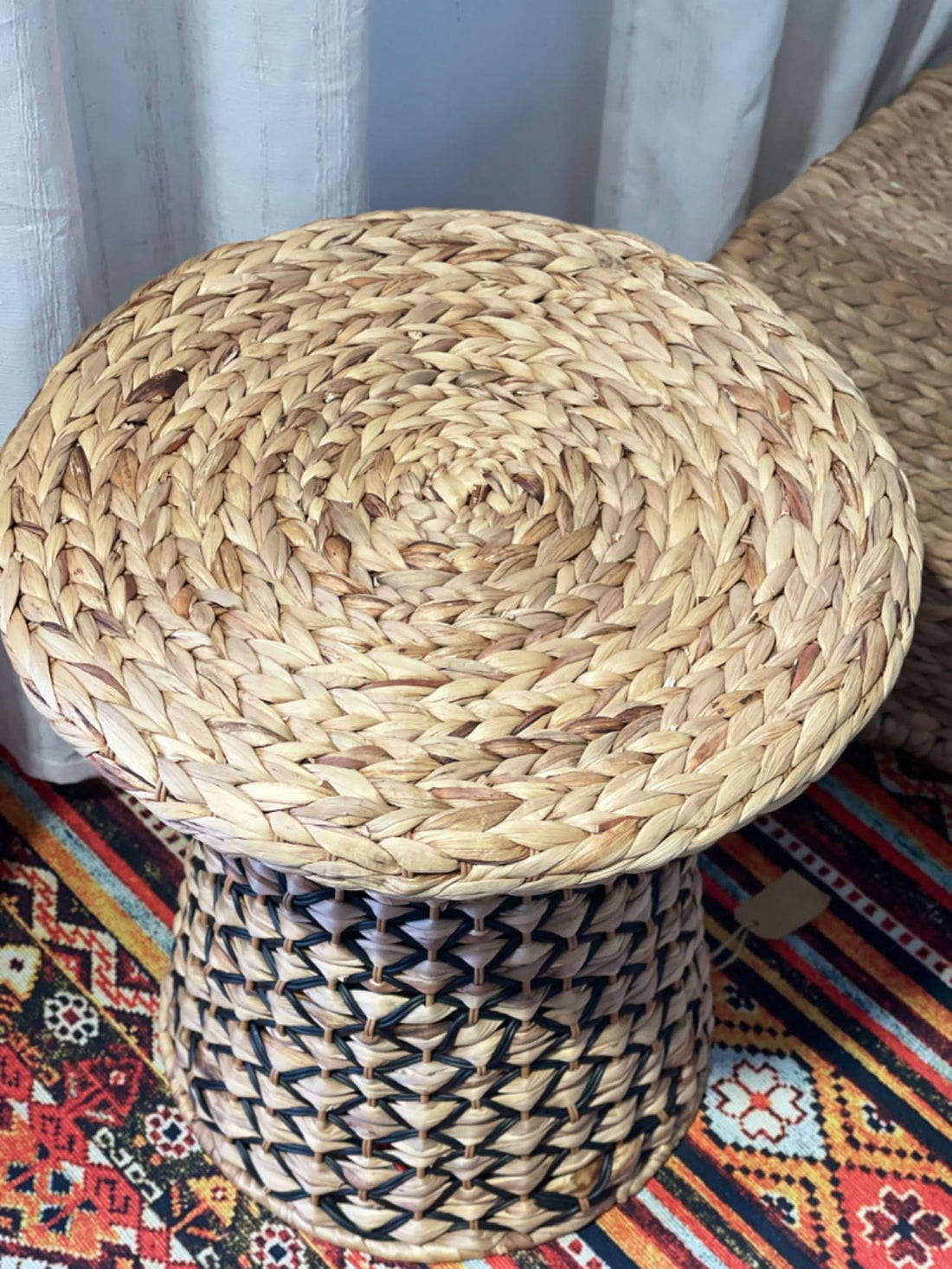 Wabi Sabi Coffee Table Straw: Round woven natural straw top with black and tan braided base on a colorful rug.