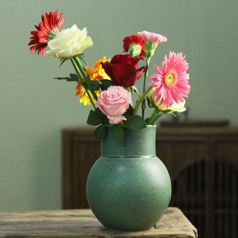 Small sage green ceramic vase holding a bouquet of colorful flowers.