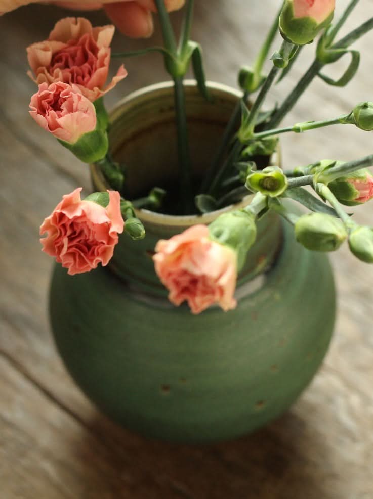 Small sage green ceramic vase holding pink carnations on a wooden surface.