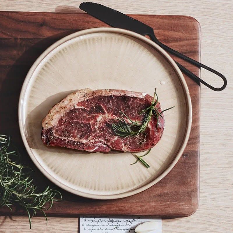 Flat ceramic glazed plate with raw steak, rosemary, and knife on wooden board.