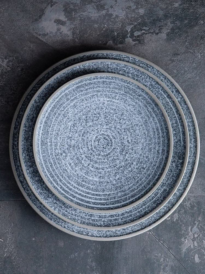 Stack of speckled stoneware flat plates with grey rims on a dark textured surface.