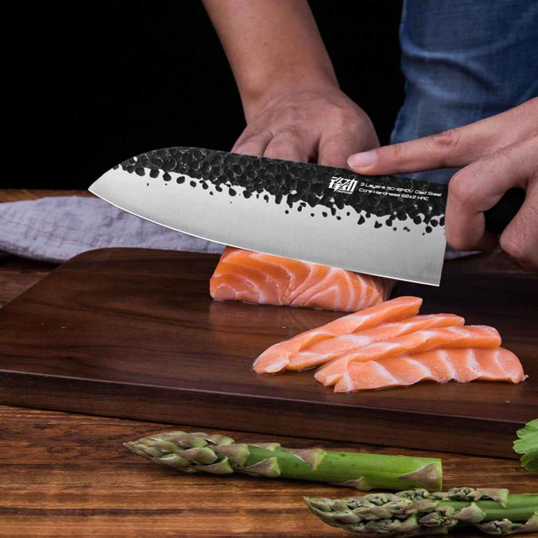 Black hammered Japanese Forged Knife slicing salmon on wood cutting board with asparagus.