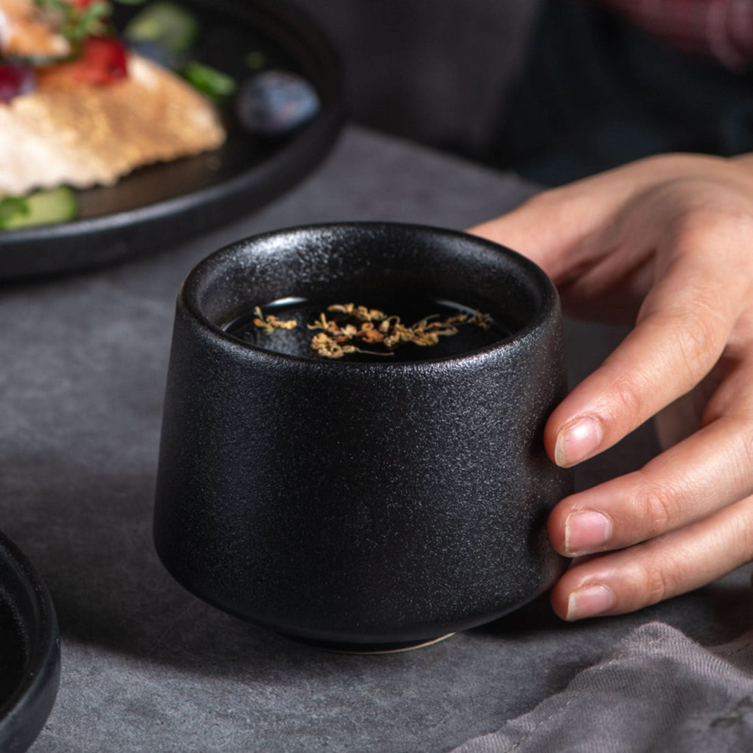 A hand holds a textured, gray-toned Japanese tea cup filled with dark tea and herbs.