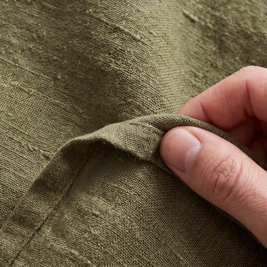 Extreme close-up of olive green linen weave and slubs with fingers pinching fabric edge showing natural texture