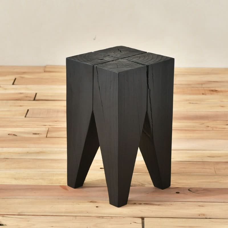 Black wood stool with faceted legs on a light wood floor.