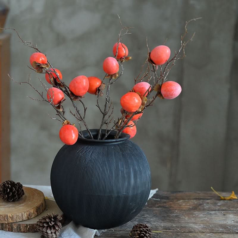 Black ceramic vase with textured ribbed finish holding faux persimmons on branches.