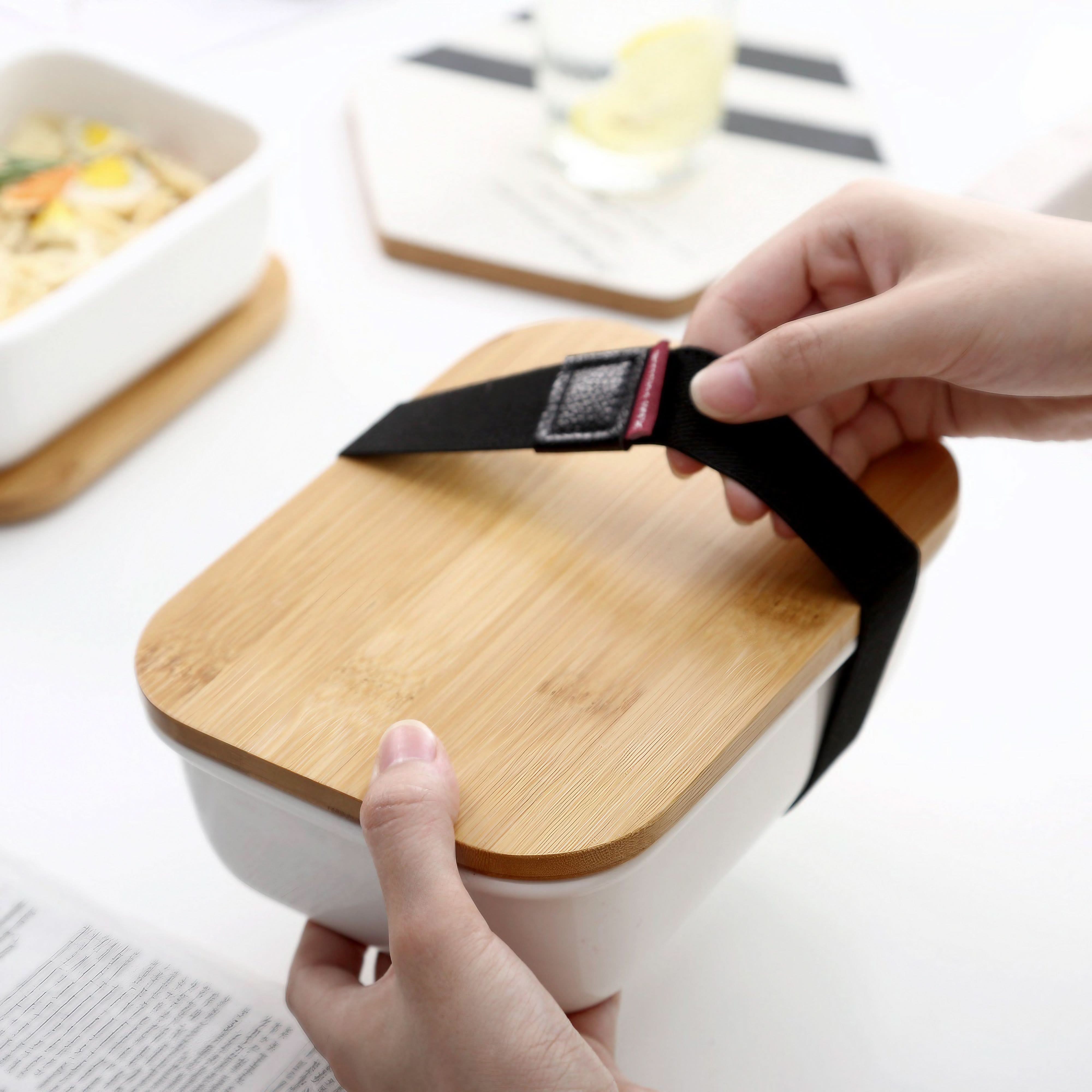 White ceramic bento box with natural wood lid and black strap, held by hands on white surface.