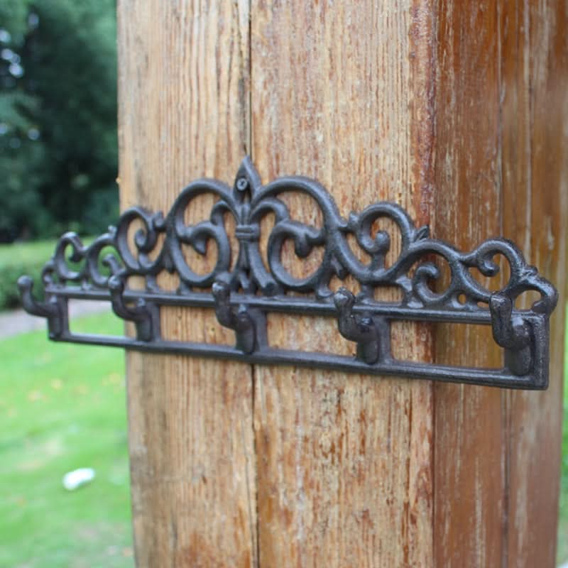Black ornate outdoor iron wall hanger with five hooks mounted on wood, in garden setting.