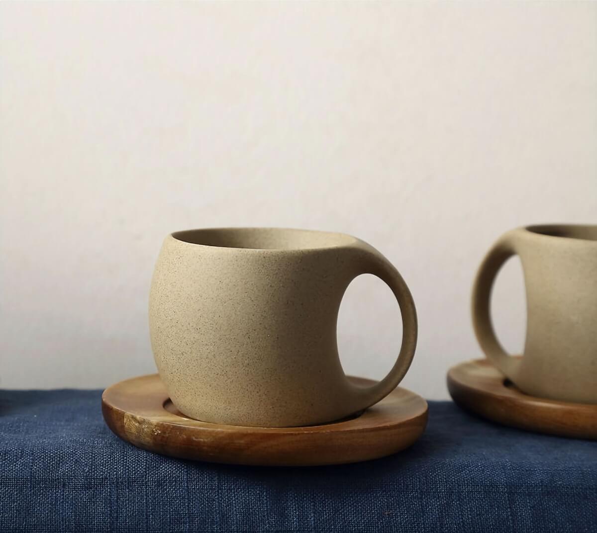 Abstract ceramic mugs with arched handle in speckled beige, on wooden saucers and blue linen.