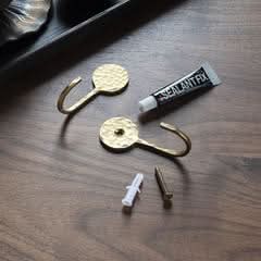 Two hammered brass wall hooks with mounting hardware and adhesive on a wooden surface.