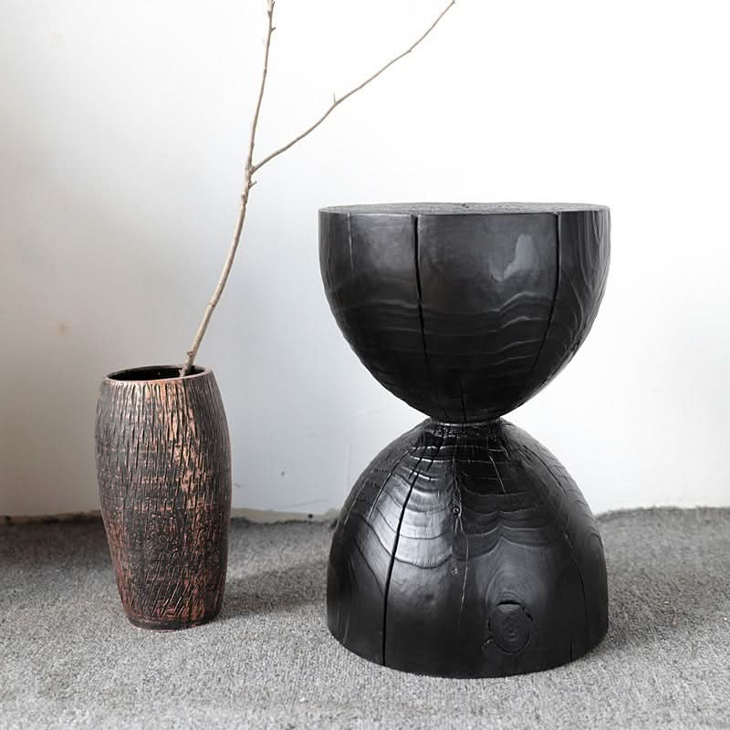 Black wood stool with textured hourglass shape on grey carpet, next to a decorative vase.
