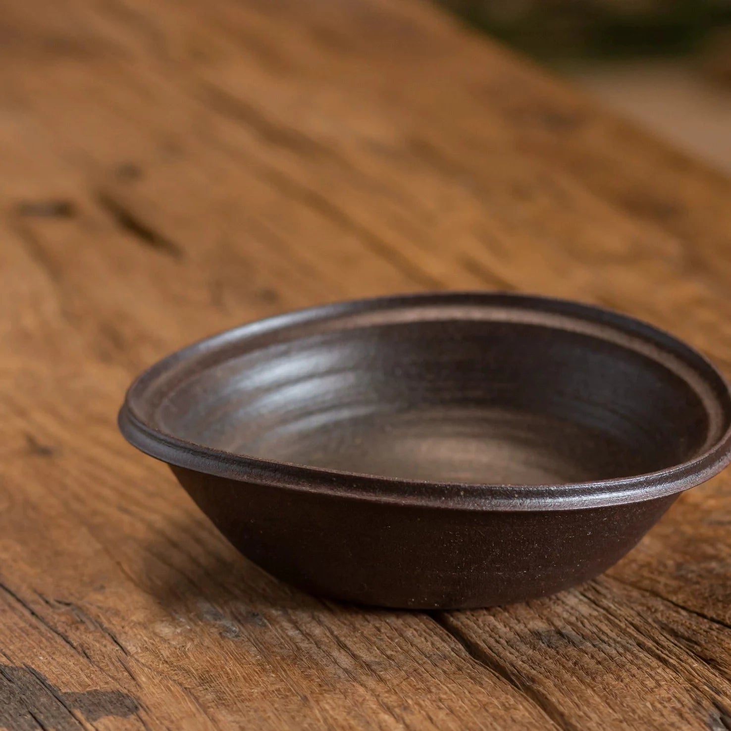 Close-up of a rustic dark brown ceramic bowl with a slightly irregular rim on a wooden surface.