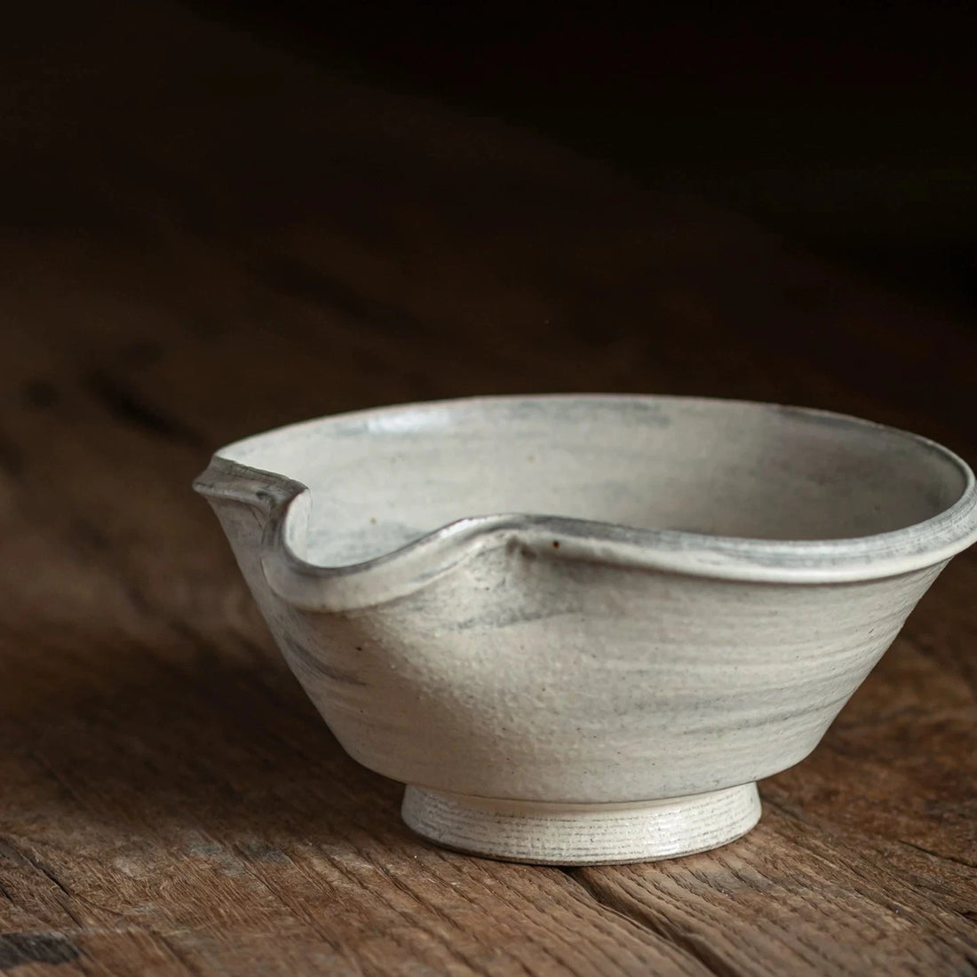 Close-up of a light grey ceramic tea bowl with an uneven, spouted rim, resting on a dark wood surface.