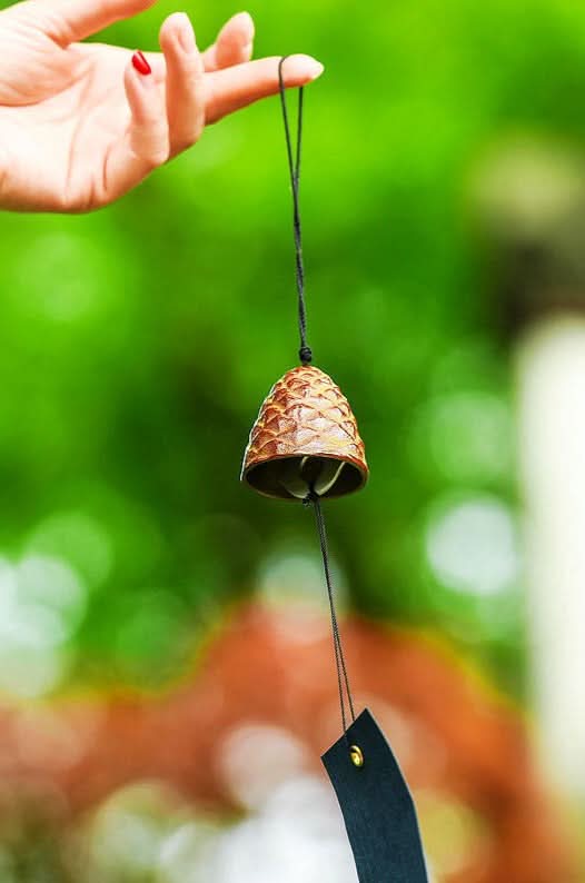 Bronze Japanese Pine Cone Wind Chime held by a hand, against a blurred green background.