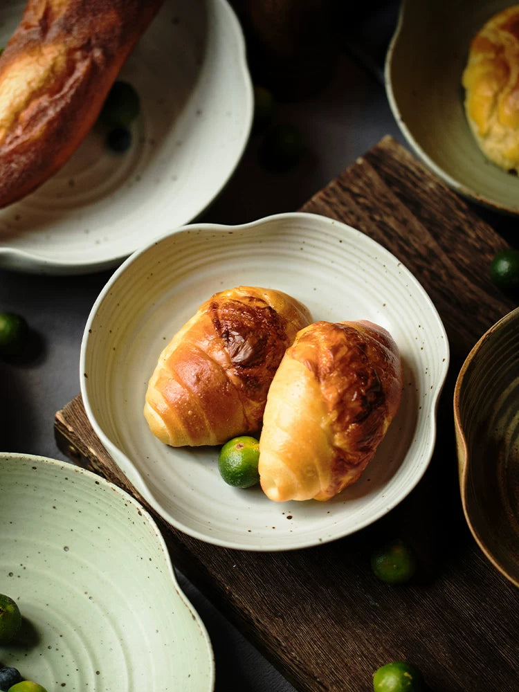 Assortment of stoneware plates with uneven edges holding croissants and bread, rustic dark setting.