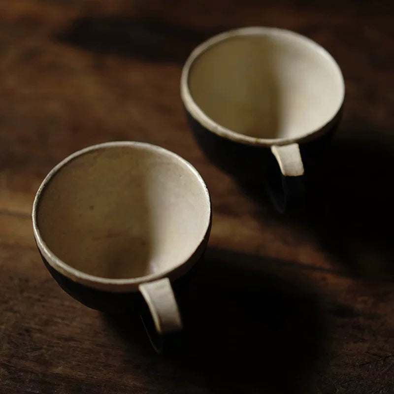 Top-down view of two artisan ceramic coffee cups with black exteriors and cream interiors resting on a dark wood surface.