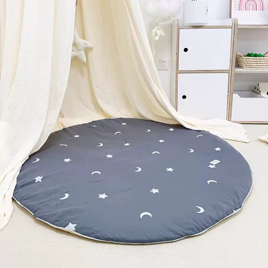 Round dark grey play mat featuring white stars and crescent moons, under a sheer curtain.
