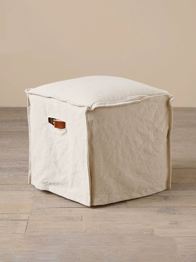 Beige linen square footstool ottoman with leather handle on light wood floor.