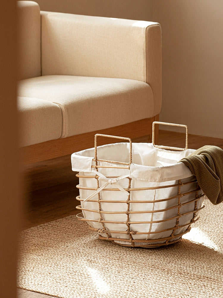 Rattan laundry basket with white linen liner on jute rug, next to neutral sofa.