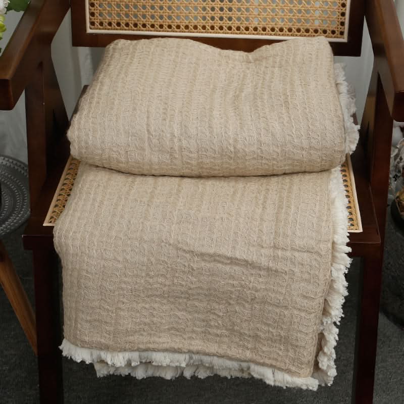 Beige linen cotton throw blanket with tassels, folded on a wooden chair.