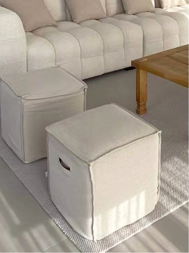 Beige linen square footstool ottoman in minimalist living room with tufted sofa and wood coffee table.