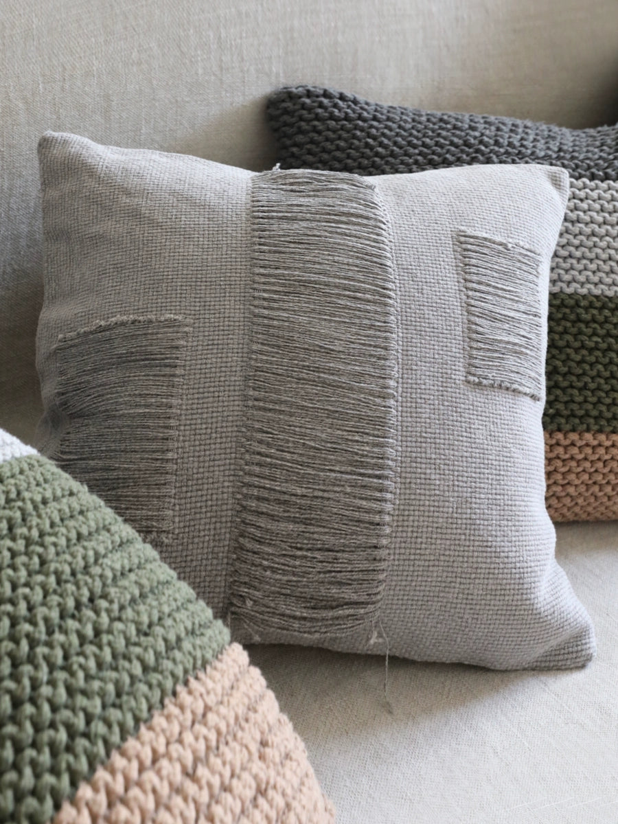 Gray woven throw pillow with fringe, part of a set of 4.  Modern boho style.  Knitted pillows in green and beige visible.