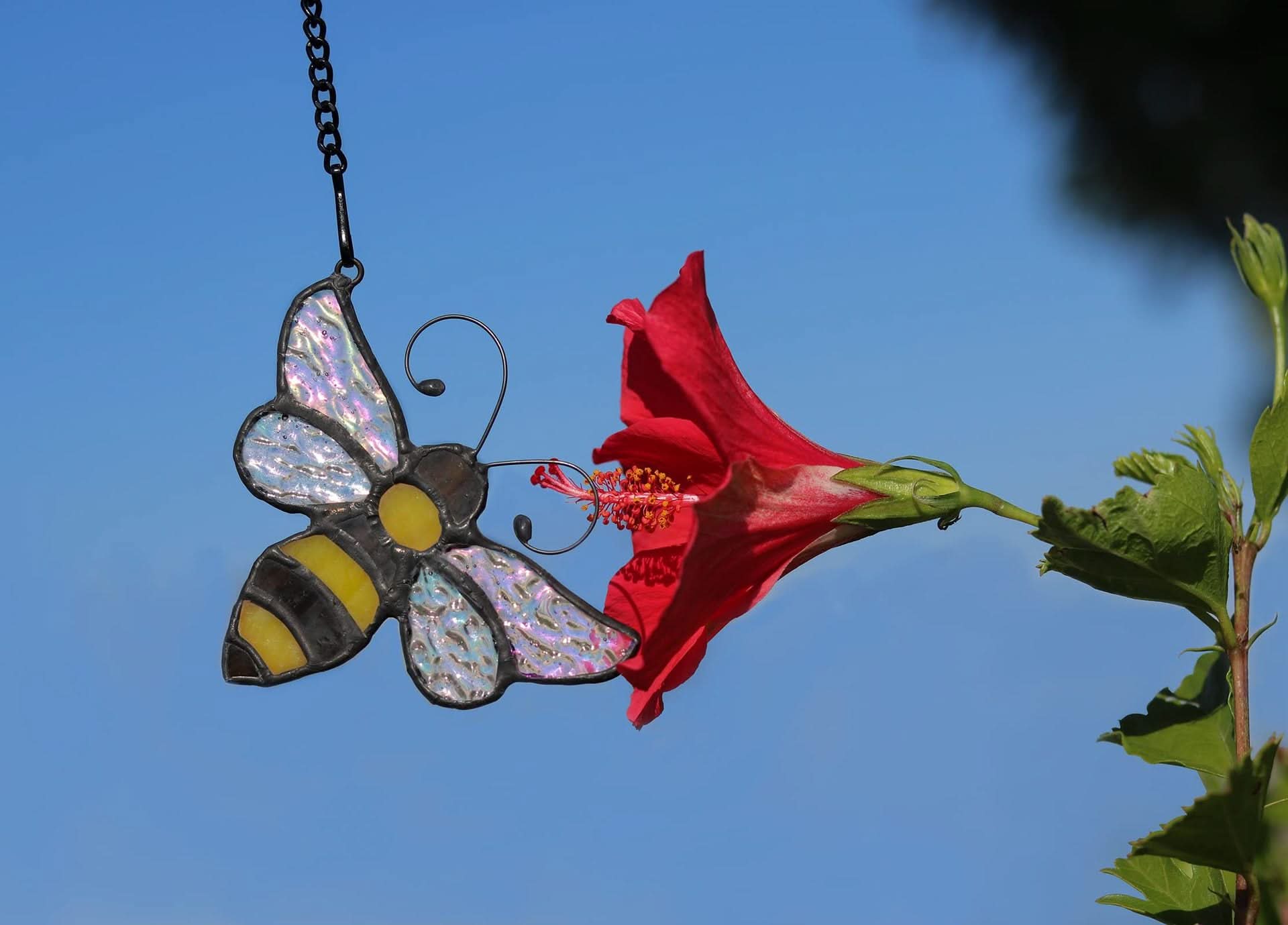 Stained glass bumblebee suncatcher with iridescent wings near a red hibiscus flower against a blue sky.