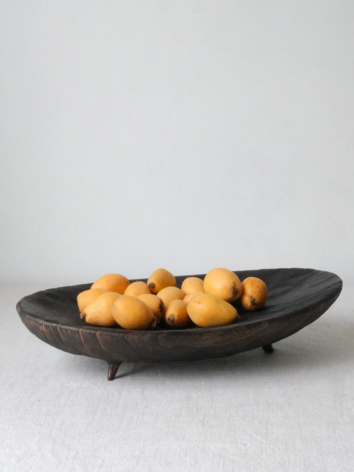 Dark brown wooden oval platter with fruit.  Small, round, yellow fruits are inside. The platter has three small dark brown feet.