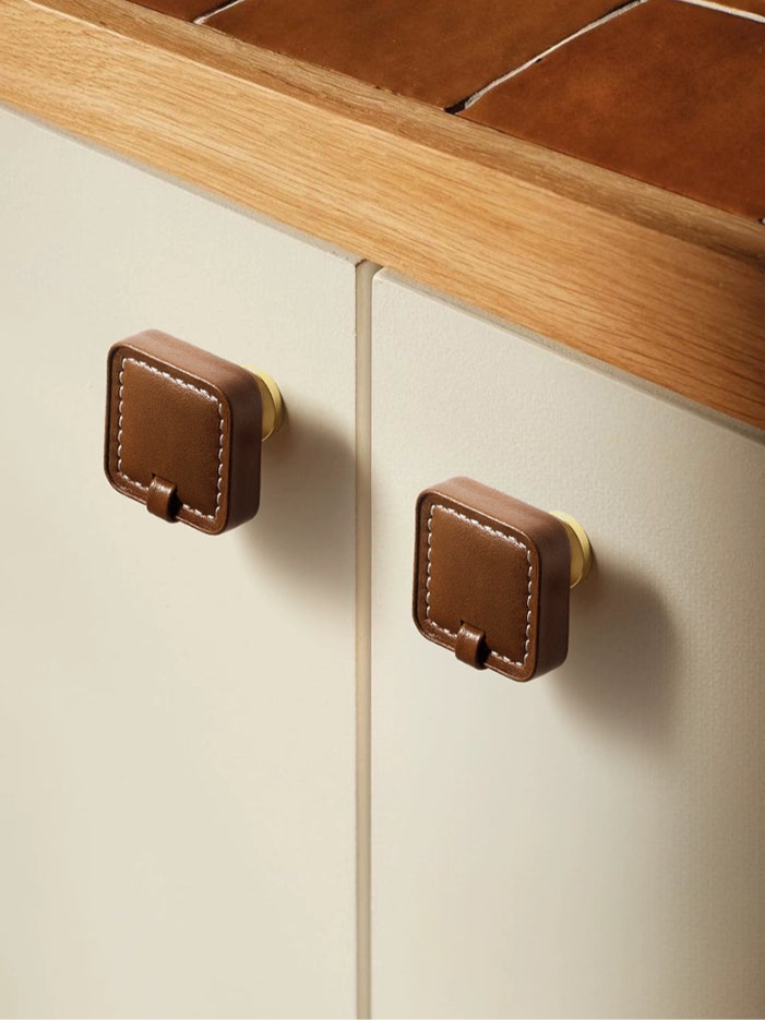 Brown leather square drawer pulls with white stitching and brass hardware on cream cabinets.