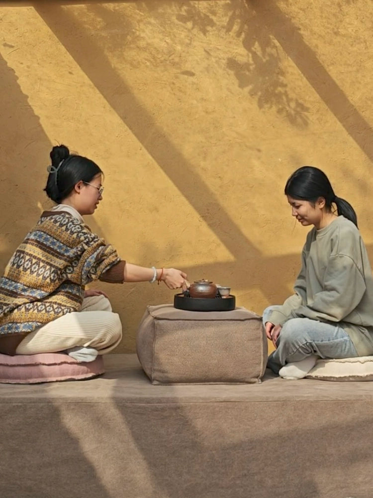 Two women seated on a light brown knitted pouf ottoman, sharing tea in a relaxed outdoor setting.  Beige and neutral tones.