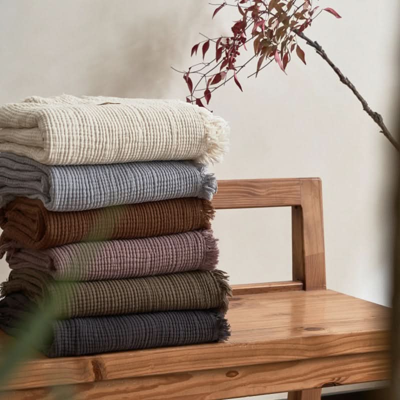 Stack of Cozy Textured Cotton Throw Blankets in muted earth tones with fringed edges on a wooden bench.