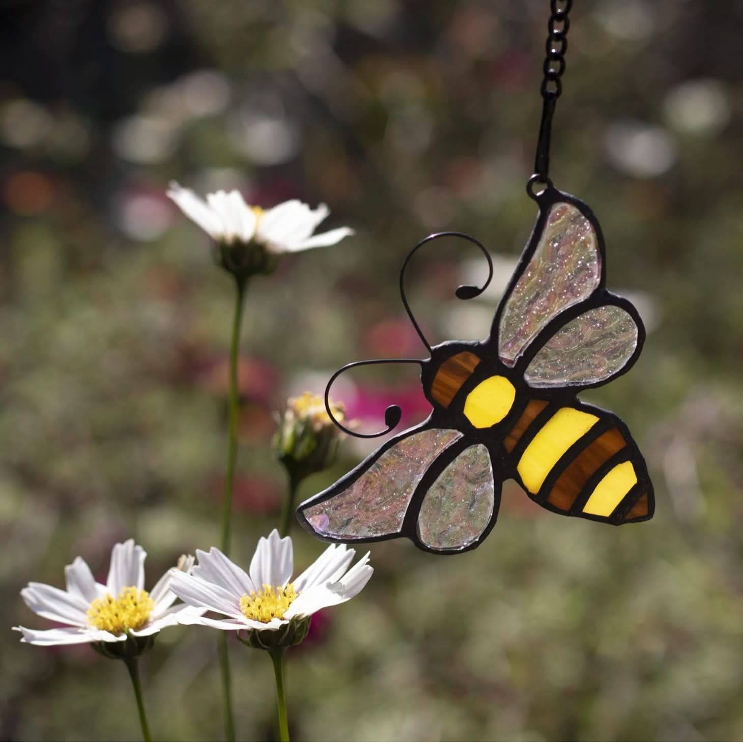 Stained glass bumblebee suncatcher with yellow & brown stripes, hanging outdoors near white daisies.
