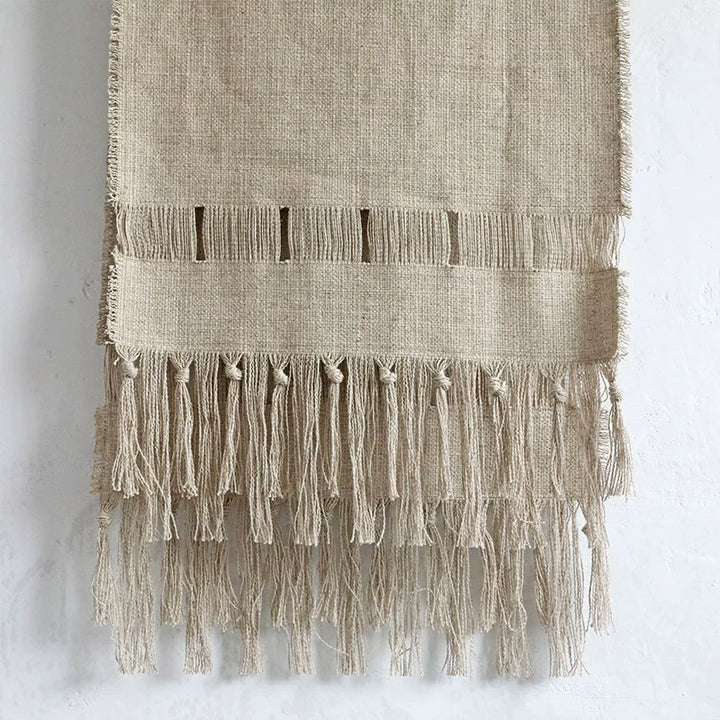 Beige linen table runner with fringe tassels. Woven design with cut-out details.