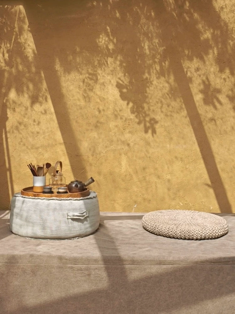Round beige fabric ottoman pouf with decorative tea set on a wooden tray, next to another pouf.