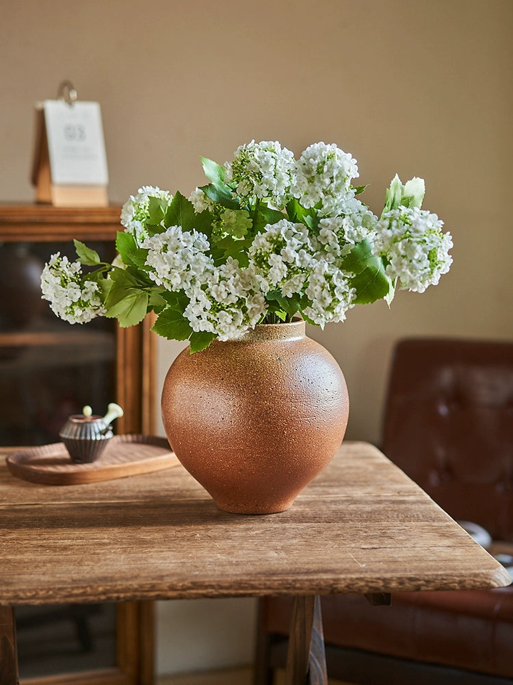 Rustic brown handmade vase, filled with white flowers, on a wooden table.  Pottery with unique texture.