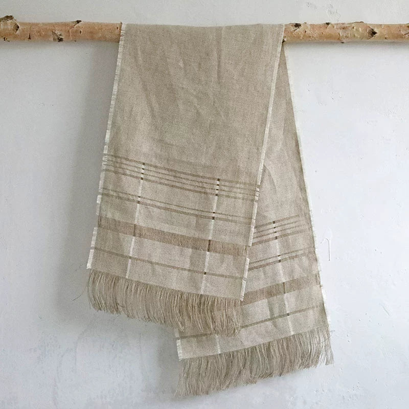 Natural beige linen table runner with fringes and subtle stripes, displayed on a branch.