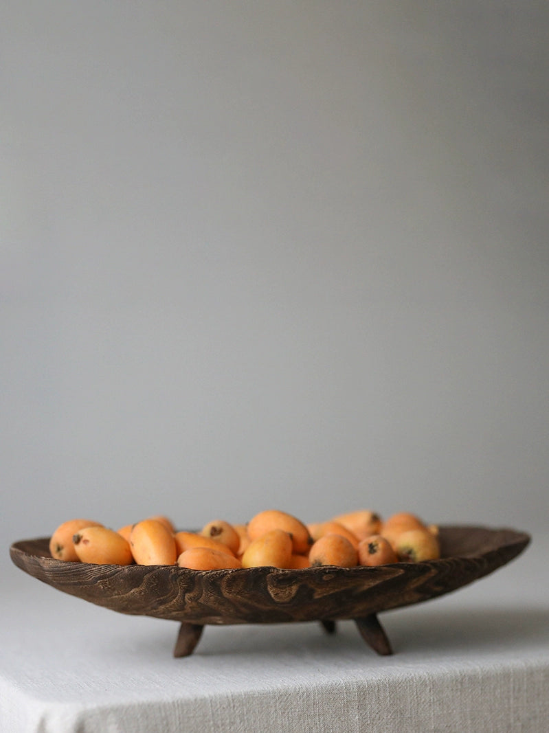 Wooden fruit bowl with small, orange-yellow fruits.  The bowl is dark brown and has a carved design, sitting on three small wooden legs.