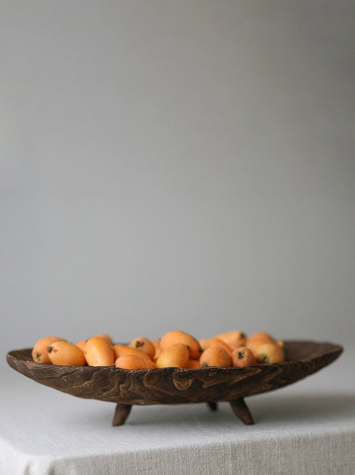 Wooden fruit bowl with small, orange-yellow fruits.  The bowl is dark brown and has a carved design, sitting on three small wooden legs.